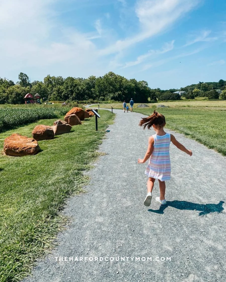 Girl in striped summer dress skips along flat, gravel trail at Poplar Grove Park in Street, Maryland. Perfect for a visit to a farm-themed playground and connects to the Darlington Library