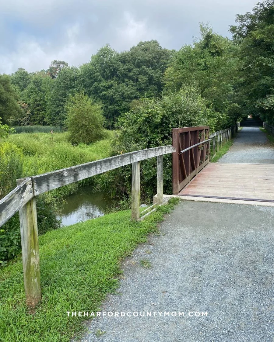 wooden bridge over narrow creek along MA & PA trails in Bel Air, Forest Hill, and Fallston, Maryland. Lush, green trees line the trail leading to Annie's Playground and Friends Park
