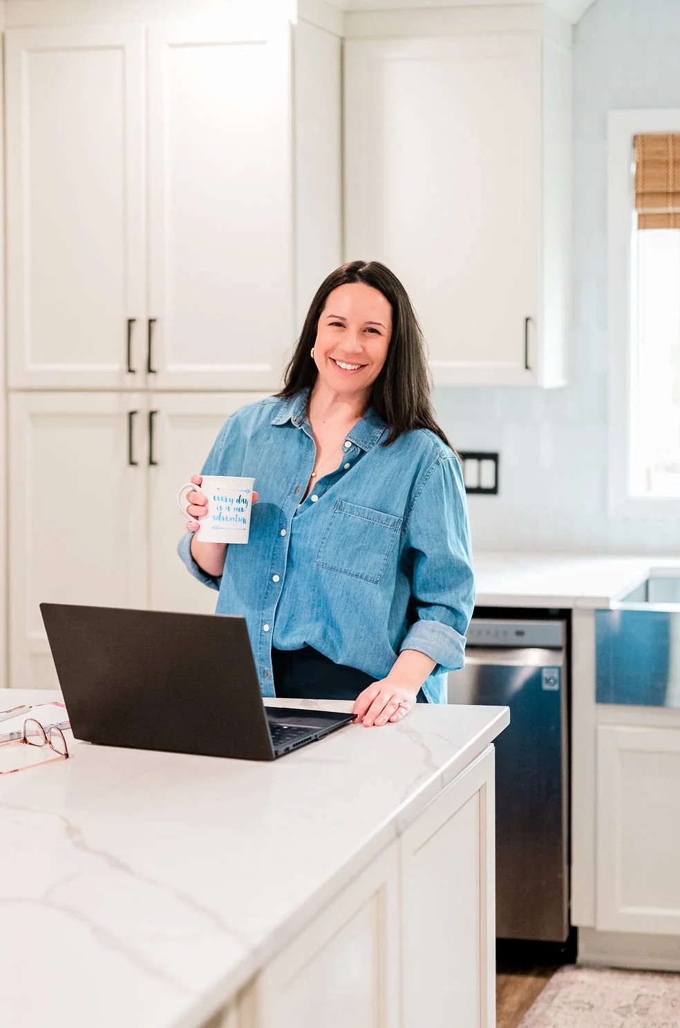 Angela stands in white kitchen wearing chambray top holding coffee mug while designing custom squarespace websites and brands on her laptop
