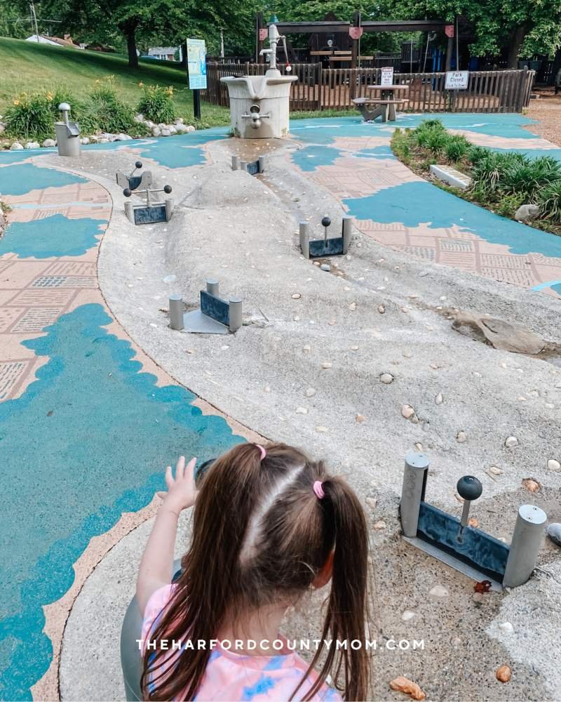 Girl plays with sandbox with splash pad in the distance at the one-of-a-kind playground at Rockfield Creative Playground in Bel Air, MD. Perfect for summer!