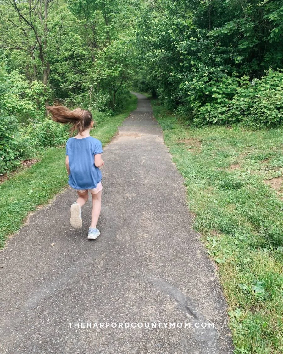 Girl in blue shirt jogs along paved trail lined with lush, green trees and shrubbery at Rockfield Park in Bel Air, Maryland