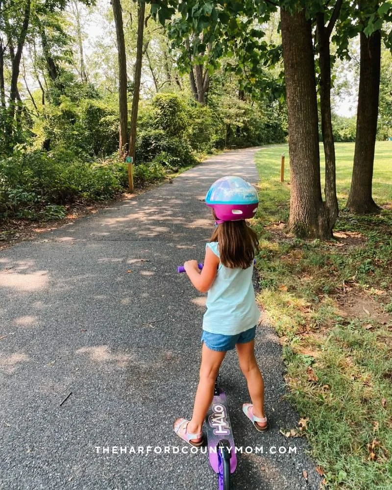 A girl in helmet stands on top of scooter on the paved, shaded Mariner Point Park trail in Joppatowne, Maryland.