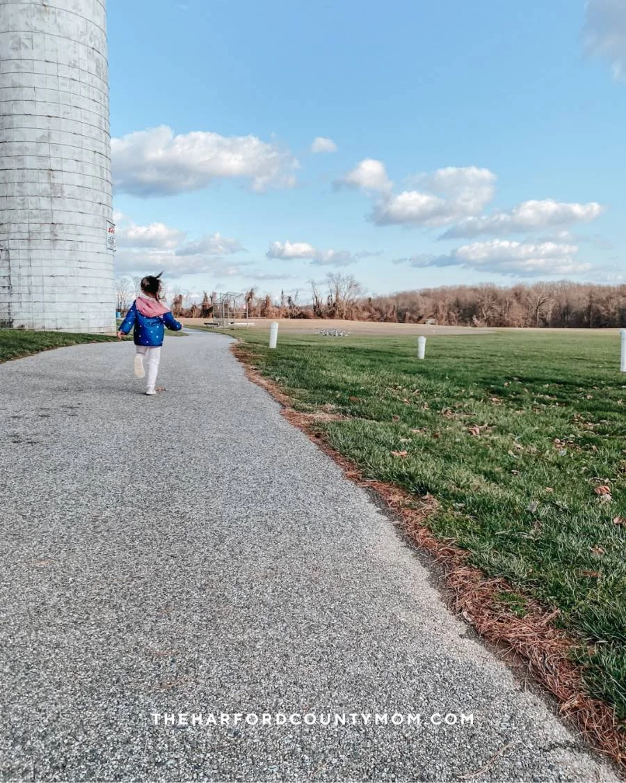 Girl in jacket runs along open, paved trail at Churchville Recreation center in Churchville Maryland with cylindrical structure on the left