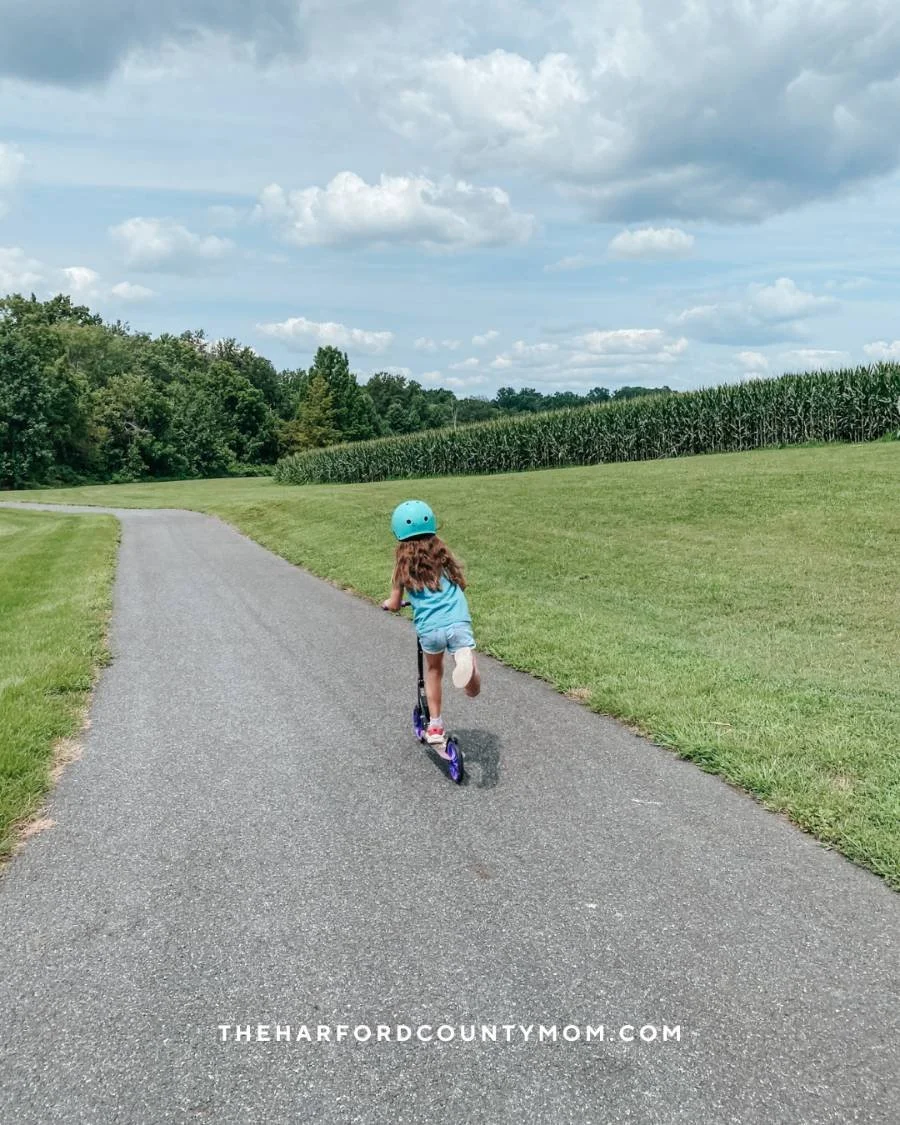 Girl in turquoise helmet rides scooter on open, paved loop trail with crop fields off to the right at Francis Silver Park in Darlington, Maryland