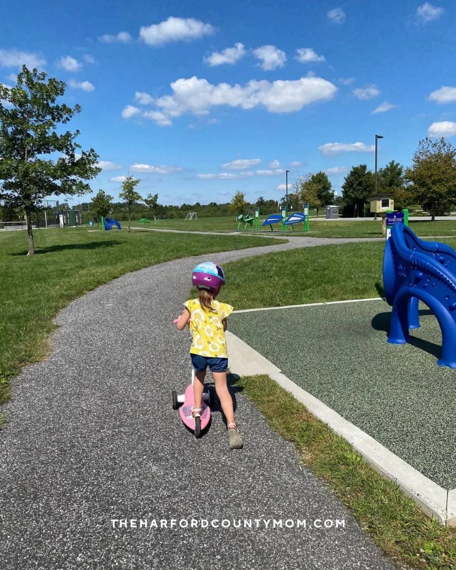 Girl in yellow top and helmet rides scooter along paved trail at Schucks Park in Bel Air, Maryland. Playground equipment is along the trail.