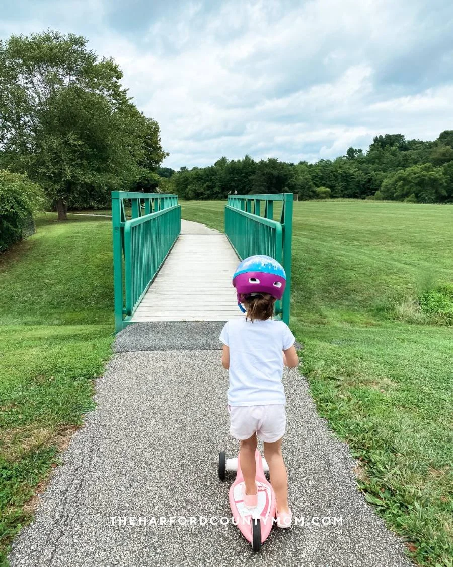 Girl in helmet rides scooter along paved loop trail at Fox Meadows Park in Jarrettsville, Maryland