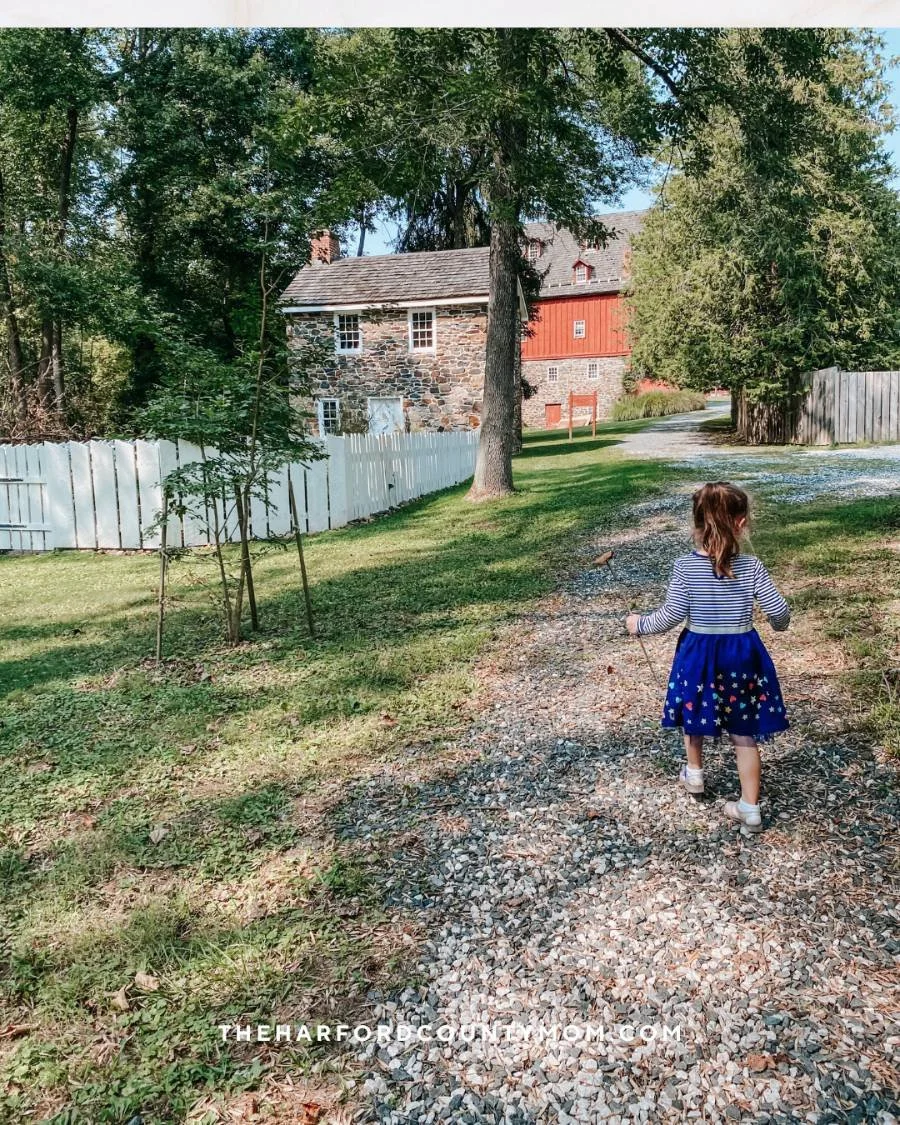 Girl in blue dress walks along gravely dirt trail with historical Jerusalem Mill in the background on the Jerusalem Mill Trail in Kingsville, Maryland. Perfect for a historical hike