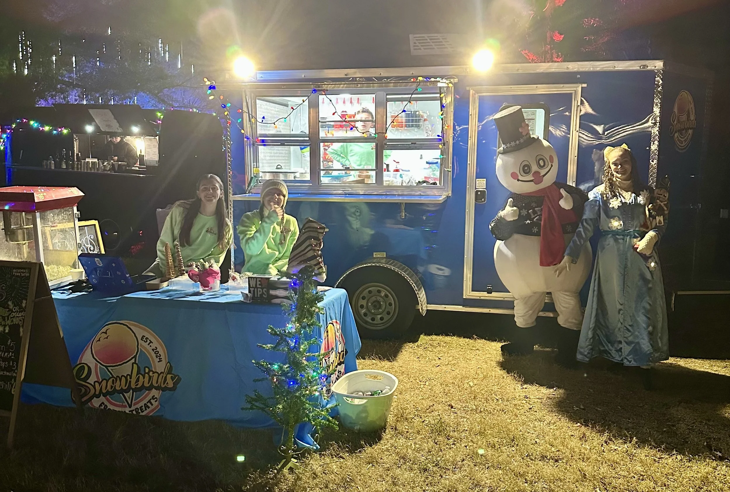 A nighttime scene of a food truck decorated with Christmas lights, with two women sitting at a table selling snowbirds treats, and a person dressed as a snowman and a person in a princess costume standing outside the truck.