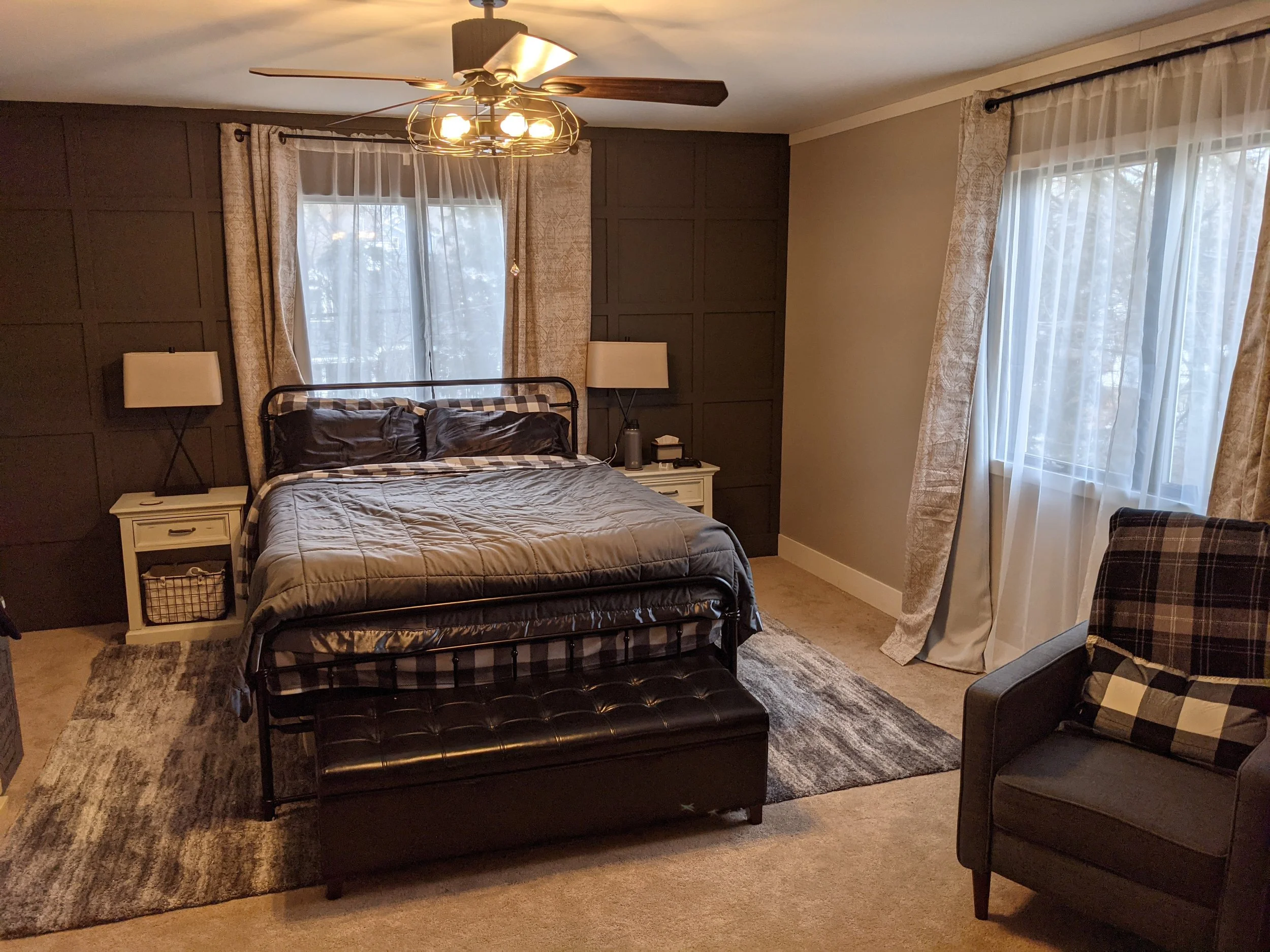 Cozy primary bedroom with black accent wall, metal bed frame, gray bedding, area rug, armchair, nightstands and neutral curtains.