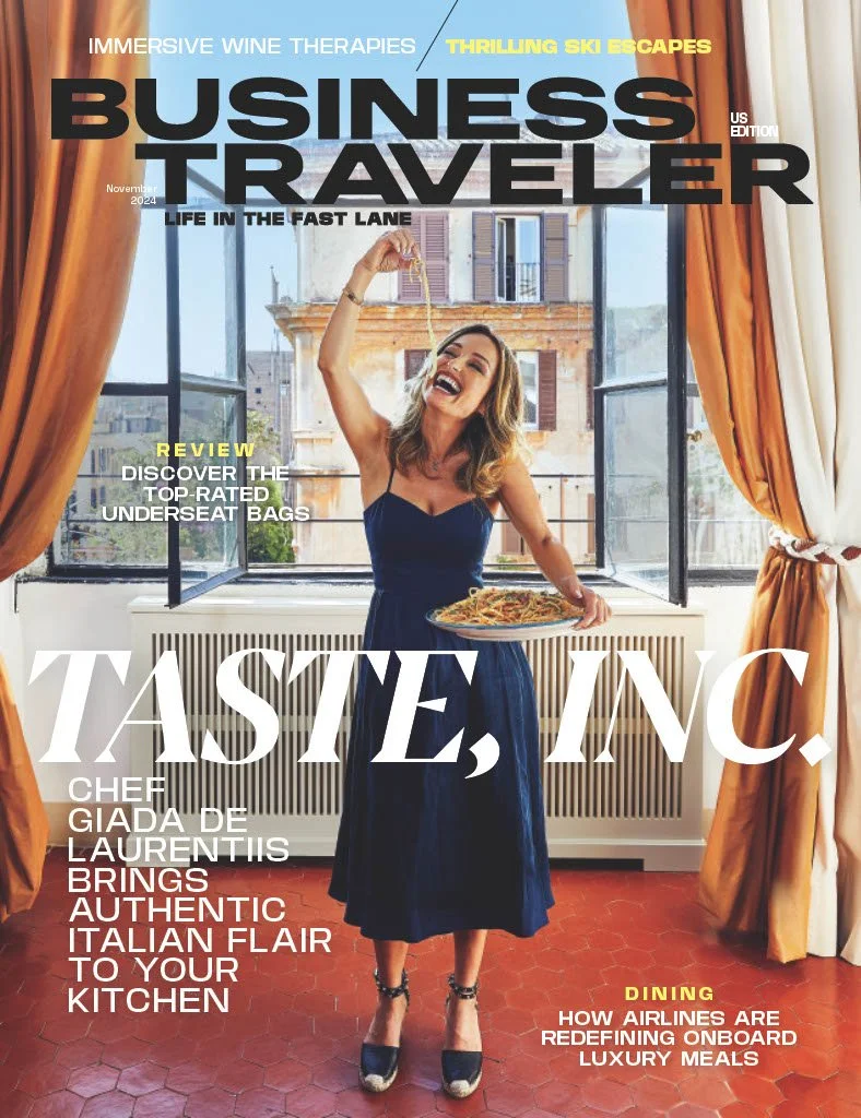 Magazine cover of Business Traveler, November 2024 issue, featuring a woman in a navy dress holding a plate of spaghetti, standing in front of a large open window with city buildings outside, gold curtains on either side, and a terracotta tile floor.