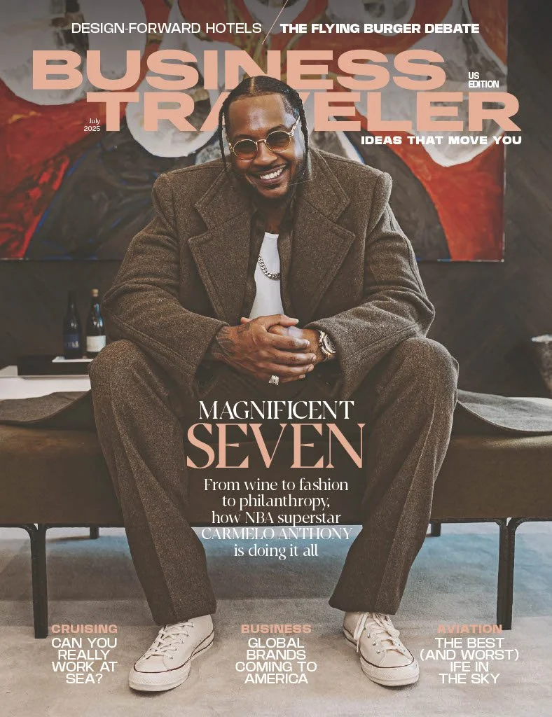 Cover of Business Traveler magazine featuring NBA superstar Carmelo Anthony sitting on a bench, dressed in a brown suit, smiling, with a large colorful painting behind him. The magazine headline highlights his achievements from wine to fashion and philanthropy.