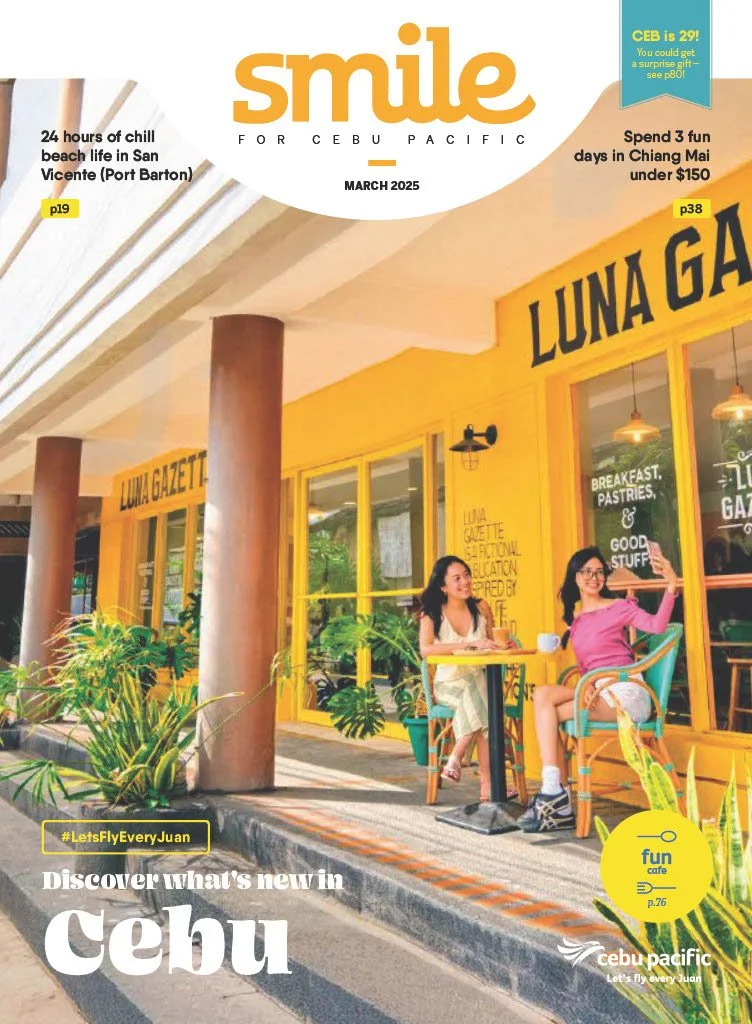 Two women sitting at an outdoor cafe table with bright yellow building labeled "LUNA GAZETTE." They are smiling and waving. Plants are in front, and the background shows a yellow restaurant and window reflections.