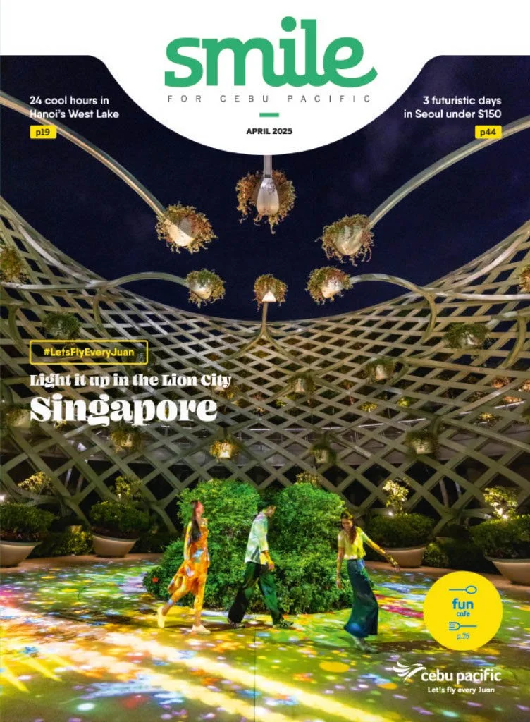 People walking in an indoor garden with a glass roof featuring hanging potted plants in Singapore, with colorful lights on the floor and a futuristic architectural structure.