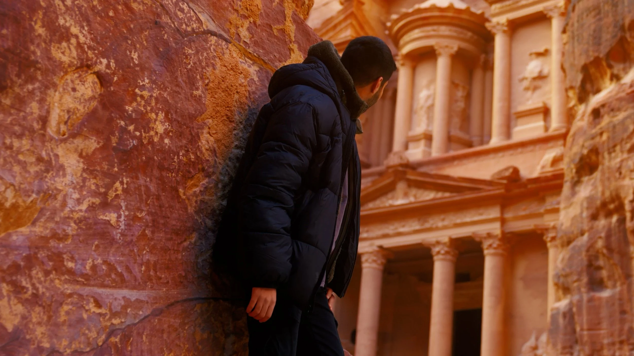 A man in a black winter jacket leaning against a large red rock formation, with the ancient city of Petra's Treasury building in the background.