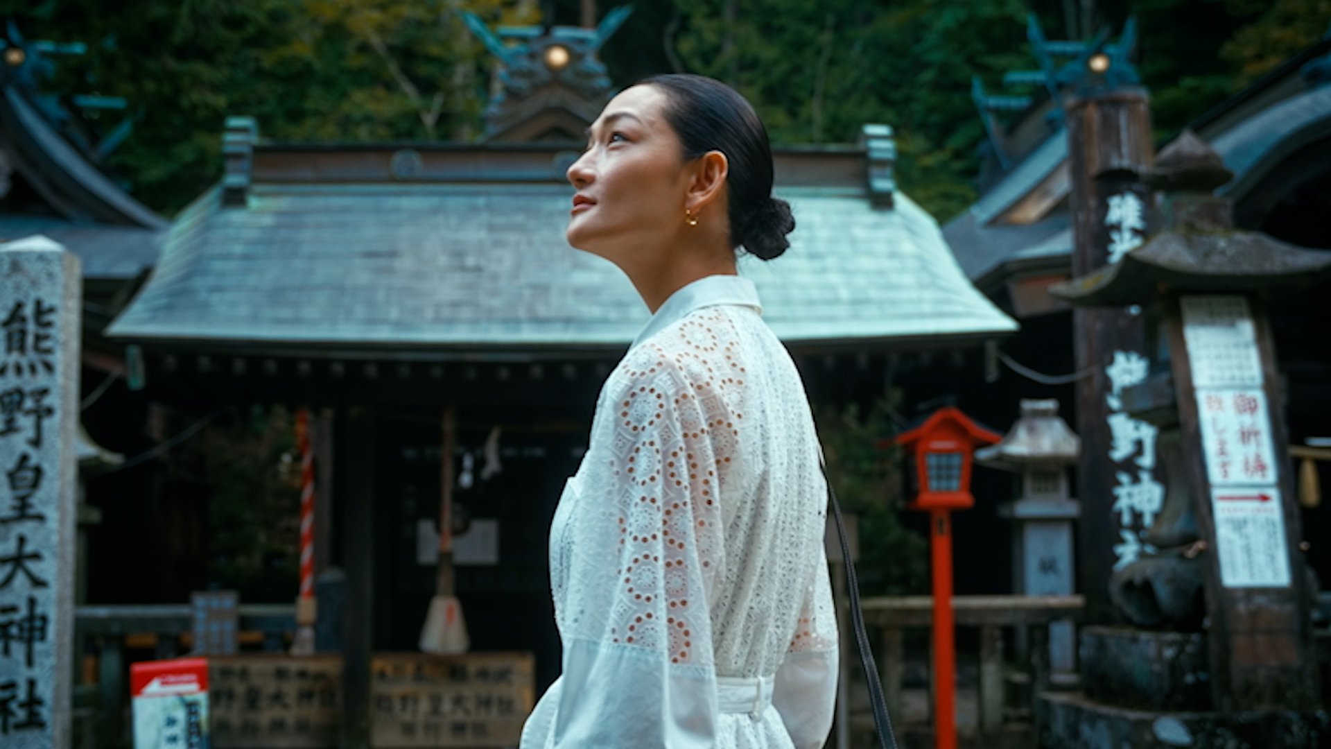 A woman wearing a white lace dress standing outdoors in front of a traditional Japanese shrine hall, with trees in the background.