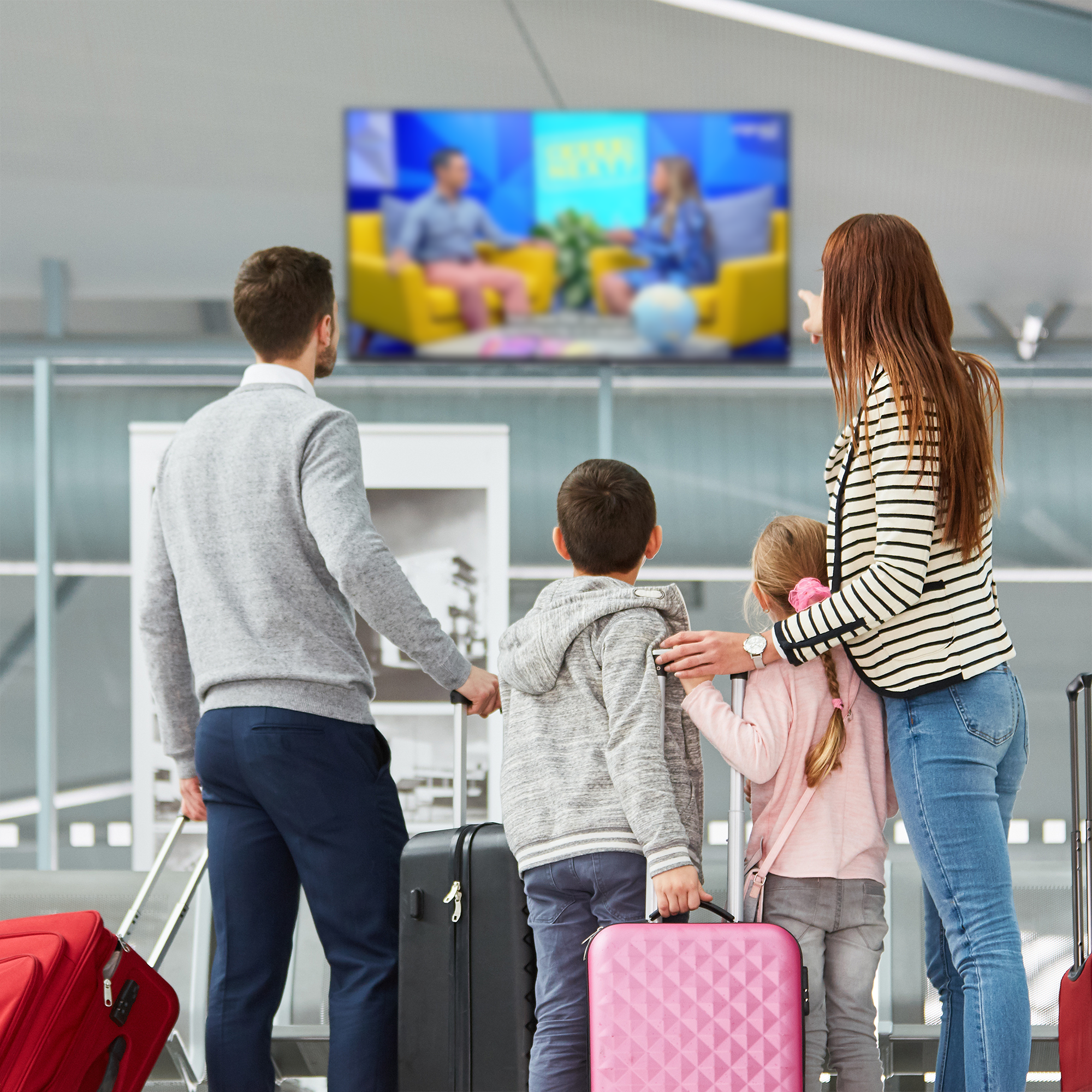 A family of four, including a man, a woman, and two children, waiting at an airport gate with their luggage, watching a television screen.
