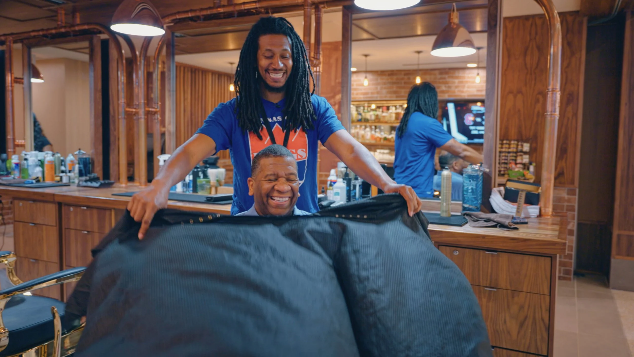 A hairstylist and a man in a barbershop, with the barber smiling and the customer smiling with eyes closed, holding a black cape around his shoulders.