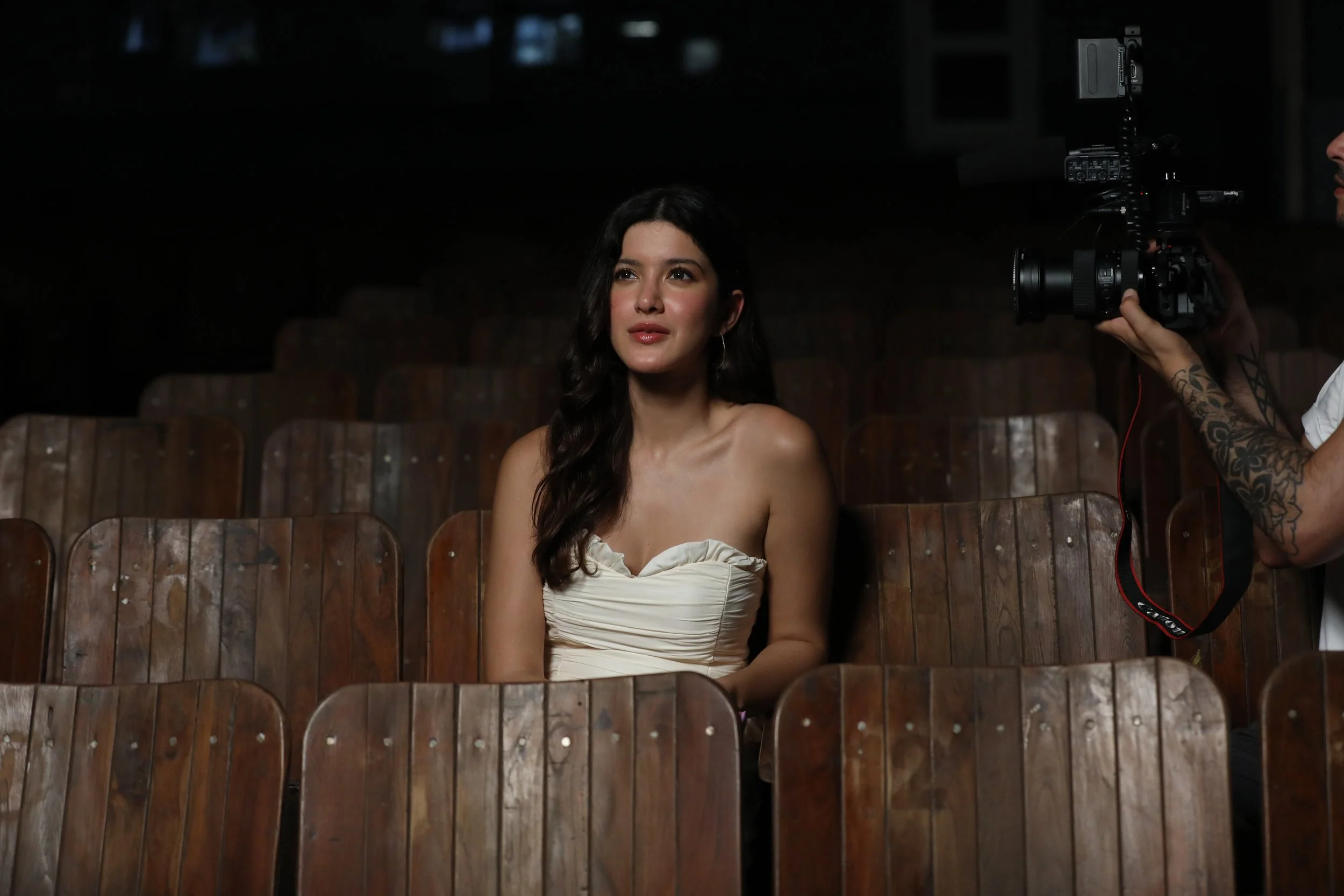A woman with long dark hair sitting in an empty theater, being filmed by a man with tattoos holding a camera.
