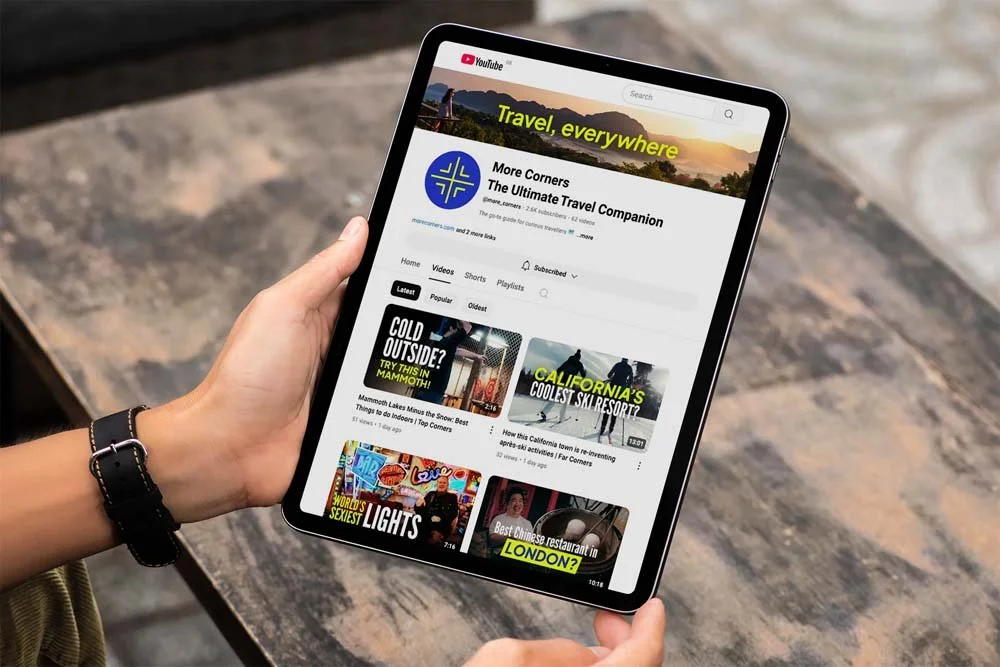 A person holding a tablet displaying a YouTube channel called 'More Corners: The Ultimate Travel Companion' with videos about travel destinations and tips.