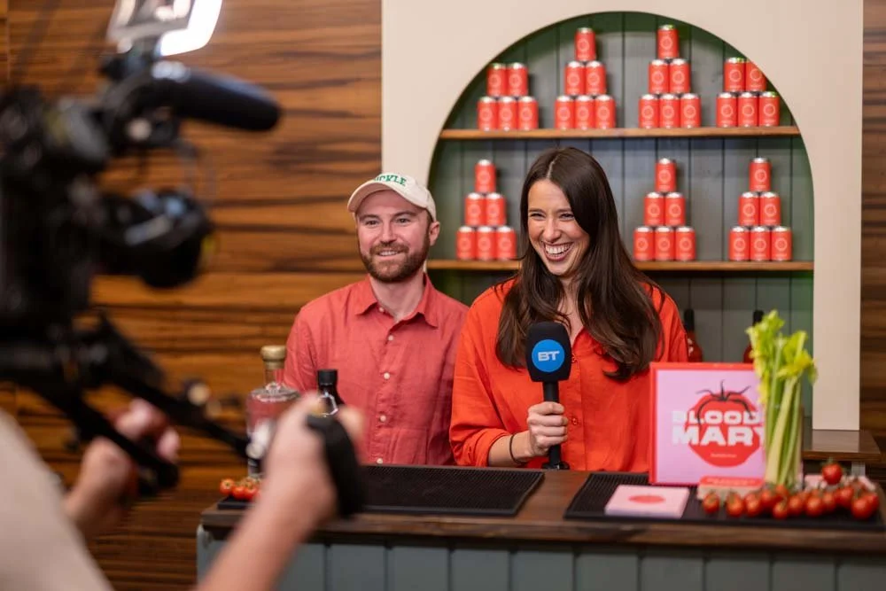 A woman holding a microphone with a BT logo, smiling, standing behind a counter with tomatoes and a sign, in a store with stacked cans on a shelf in the background. A man in a baseball cap stands beside her.