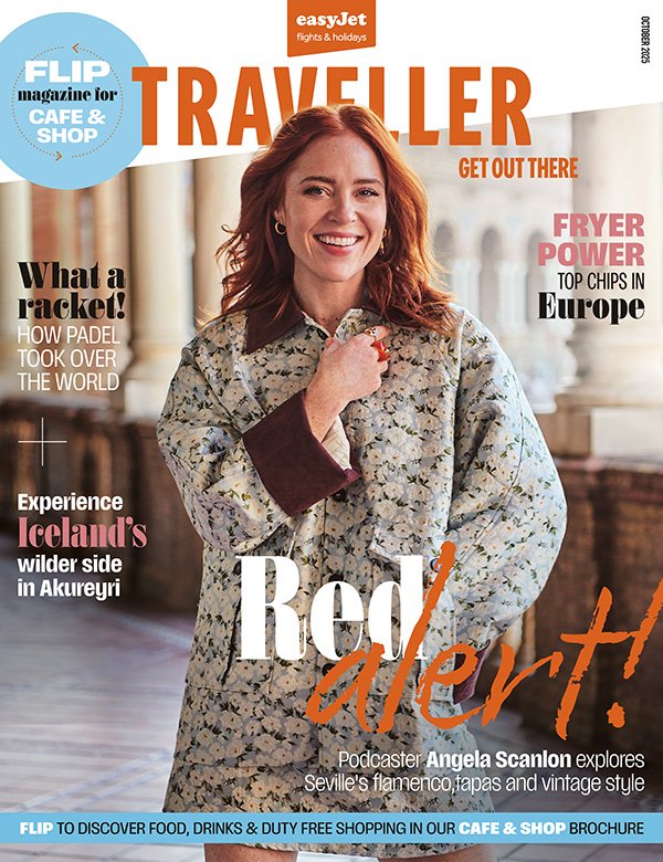 A smiling woman with red hair wearing a floral jacket with maroon cuffs on the sleeves, standing in an outdoor historic architectural setting, on the cover of a travel magazine.