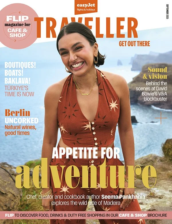 smiling woman standing outdoors near the ocean, wearing a rust-colored dress with star patterns, in front of rocks and water with cliffs in the distance.