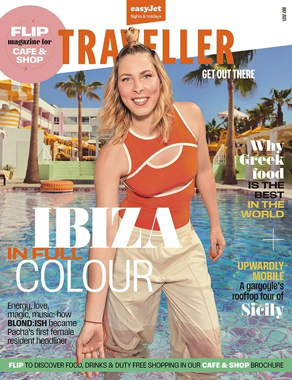 A magazine cover featuring a smiling woman in an orange and white swimsuit standing in a swimming pool at a resort with palm trees and colorful buildings in the background. The magazine is titled 'TRAILER' and has various headlines about Ibiza, Greek food, and travel.