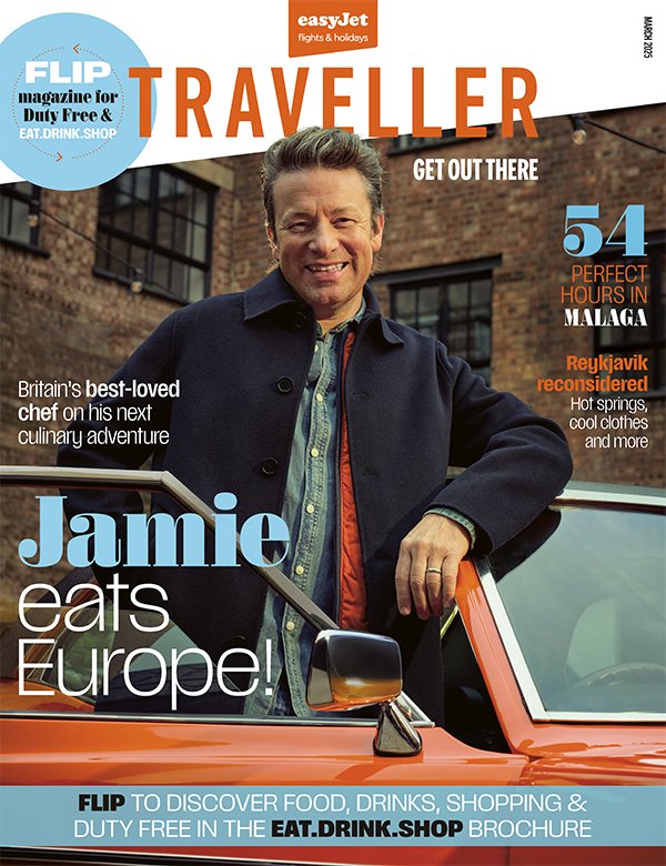 Cover of TRAVELLER magazine featuring a man smiling, standing next to a vintage orange car, with text highlighting travel features in Malaga, food, drinks, and Europe.