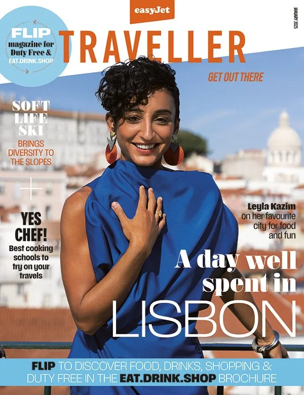 The cover of a magazine titled "Traveller" features a smiling woman with short curly hair, wearing large red earrings and a blue outfit, standing outdoors in Lisbon, Portugal, with historical buildings and a dome visible in the background.