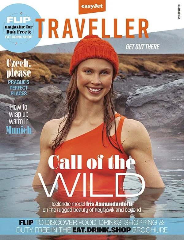 Magazine cover of 'Traveller' featuring a smiling young woman in an orange dress and knit hat, standing in a hot spring with rocky landscape in the background. The cover highlights topics like Iceland's rugged beauty, Prague's perfect places, and tips for warm-up in Munich.