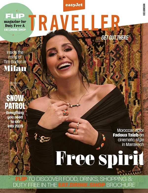Magazine cover of easyJet Traveler featuring a smiling woman with dark hair and gold jewelry, standing in front of a colorful tapestry background. The cover has headlines about travel topics and a promotional message encouraging readers to discover food, drinks, shopping, and duty-free shopping.