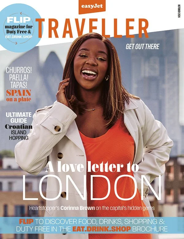Magazine cover titled "TRAVELLER" featuring a smiling woman with shoulder-length brown hair, wearing a white jacket over an orange shirt. Background shows a cityscape with tall buildings. Cover highlights travel topics like Spain, Croatia, and island hopping, and promotes a guide to duty-free shopping.