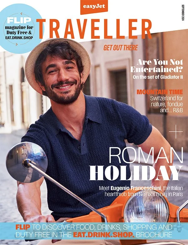 Cover of a travel magazine featuring a smiling young man with a beard, wearing a straw hat and navy shirt, sitting on a scooter in a city street. The magazine is titled 'TRAVELLER' and includes headlines about entertainment, mountain destinations in Switzerland, and an interview with actor Eugenio Franceschini.