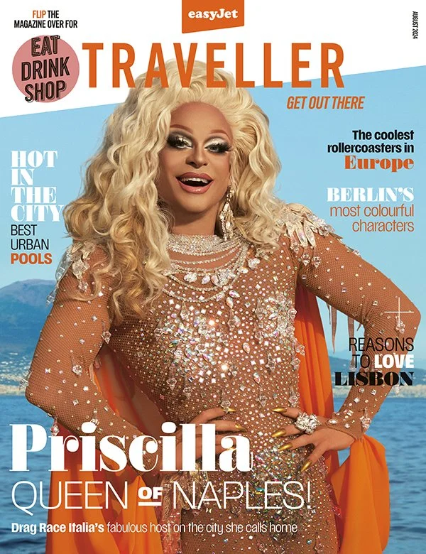 Cover of Traveller magazine featuring Priscilla, dressed in a sparkling, embellished dress with long blonde curly hair, standing in front of a water body with mountains in the background.