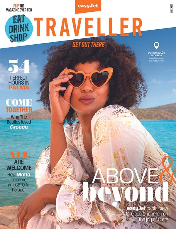 A woman with curly hair wearing orange sunglasses and a patterned light-colored dress, sitting outdoors by the water with a clear blue sky behind her on the magazine cover titled 'Traveler'.