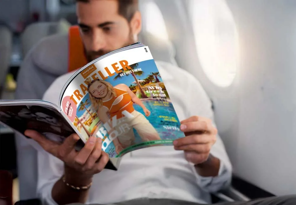A man sitting on an airplane reading a travel magazine featuring a woman in a swimsuit by a pool with palm trees.