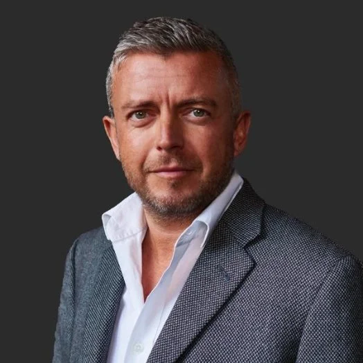 A middle-aged man wearing a gray blazer and white shirt, posing against a dark background.