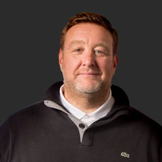 Portrait of a smiling man with short light brown hair and a beard, wearing a black Lacoste sweatshirt with a white collared shirt underneath, against a dark background.