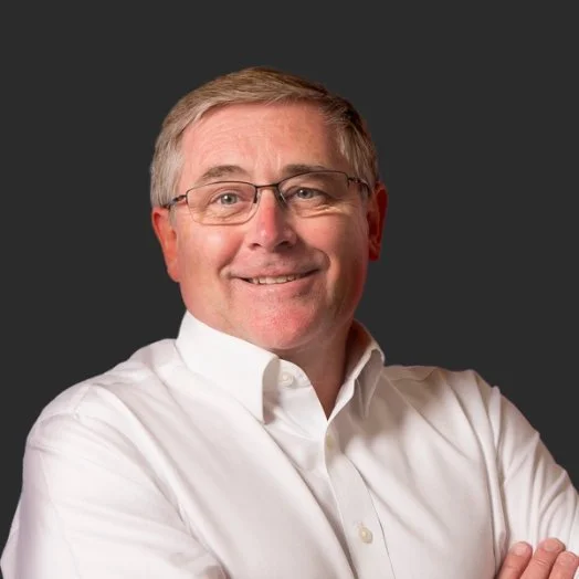 A smiling middle-aged man with glasses and gray hair wearing a white dress shirt, standing against a dark background.
