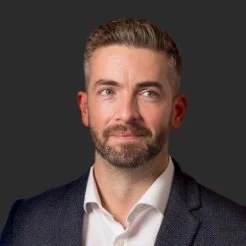 Portrait of a man with short hair and beard, wearing a suit jacket and white shirt, against a dark background.