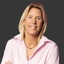 A woman with blonde hair wearing a light pink button-up shirt and a necklace, smiling with arms crossed against a dark background.