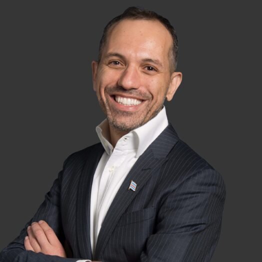 A smiling man in a dark pinstripe suit with a white collared shirt, standing against a gray background.