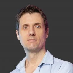 Portrait of a man with short dark hair wearing a light blue shirt, against a dark background.