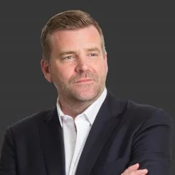 A man with short brown hair and a beard, wearing a dark suit jacket and white shirt, looking to the side with arms crossed, against a gray background.