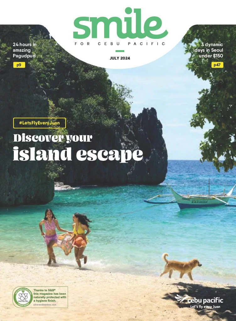 A scenic beach scene featuring two girls playing with a towel, a dog on the sandy shore, a boat floating in clear turquoise water, lush greenery, and rocky cliffs in the background, with magazine cover text promoting travel to a Cebu island in July 2024.