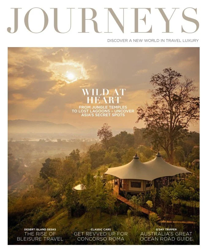 Travel magazine cover titled "JOURNEYS" showcasing a scenic landscape with a sky at sunset, lush trees, and a hut with a large, tent-like roof.