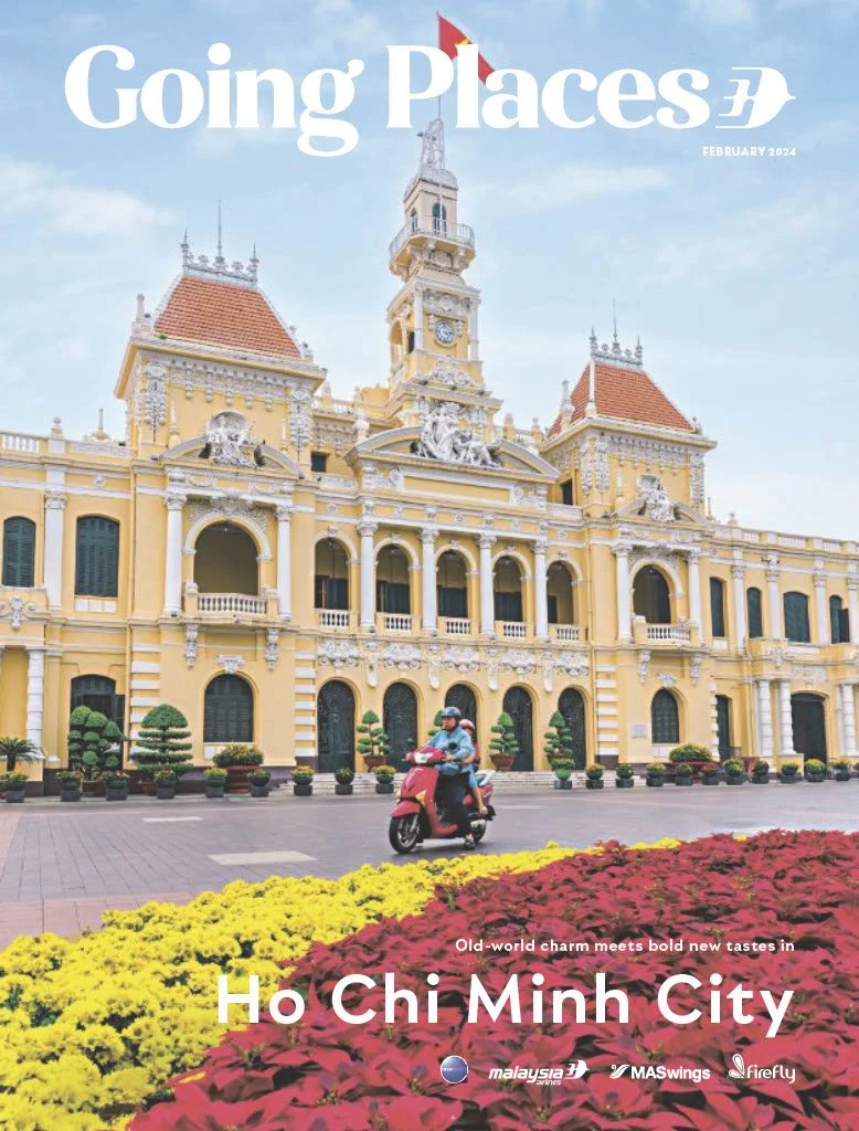 An ornate yellow and white colonial-style building, with a clock tower, situated behind a street with a person riding a pink scooter. Colorful flower beds in yellow and red are in the foreground.