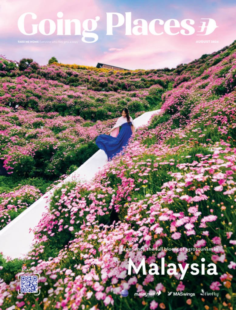 A young woman in a blue dress standing on a white slide surrounded by a hillside covered in pink and white blossoms. The sky is pink and purple with some clouds, and a building is visible at the top of the hill. The text on the image reads 'Going Places,' 'Malaysia,' and includes promotional details for agrotourism in Malaysia.