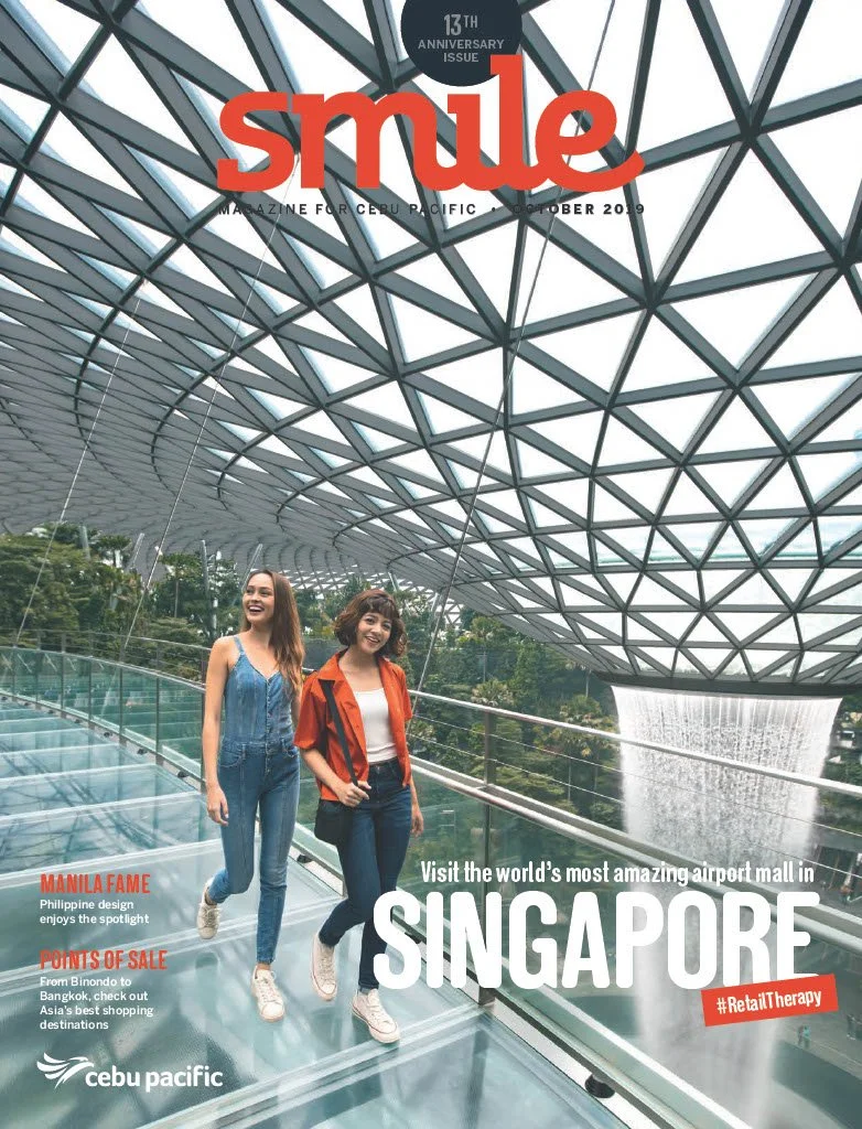 Cover of Smile magazine featuring two women walking on a glass bridge inside an airport terminal with a distinctive geometric ceiling and waterfall in the background, celebrating Singapore's airport mall.