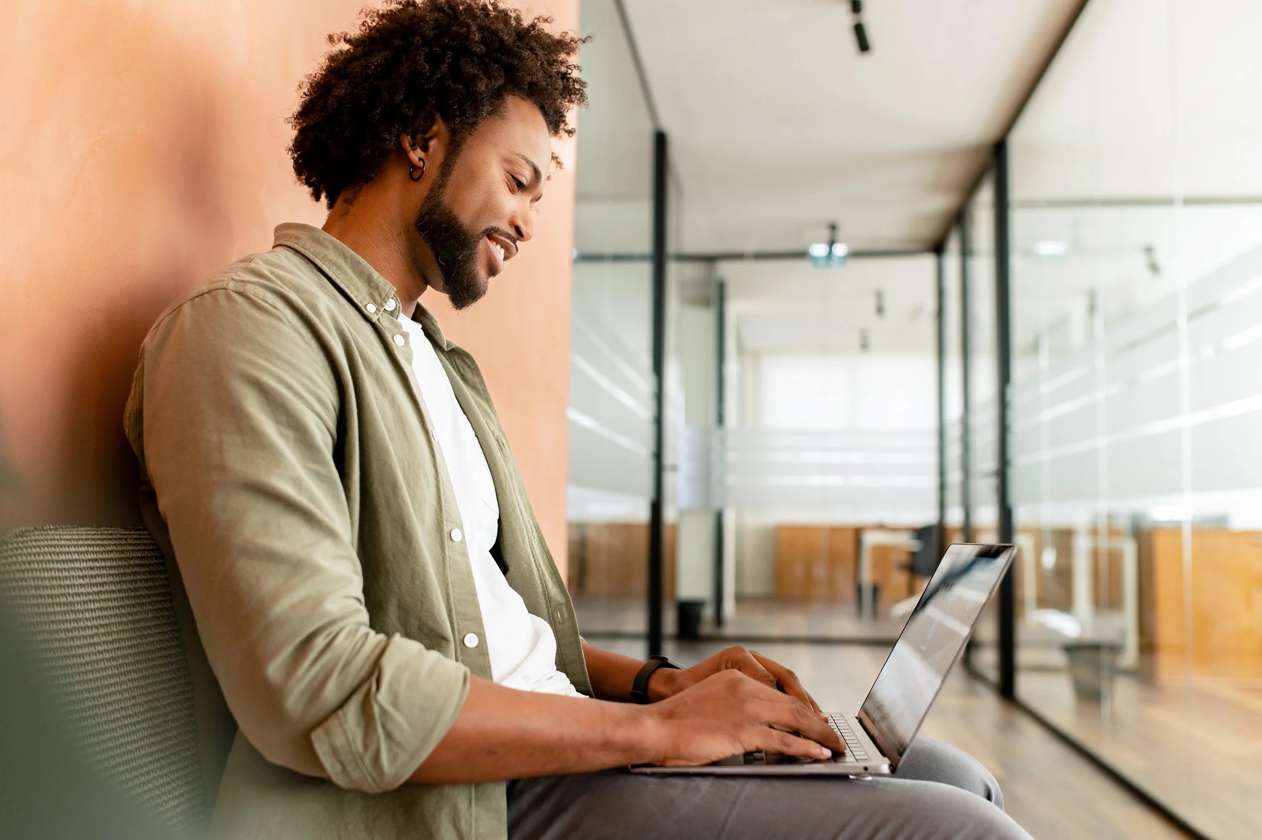 A man with curly hair and a beard sitting on a bench and working on a laptop in a modern indoor space.